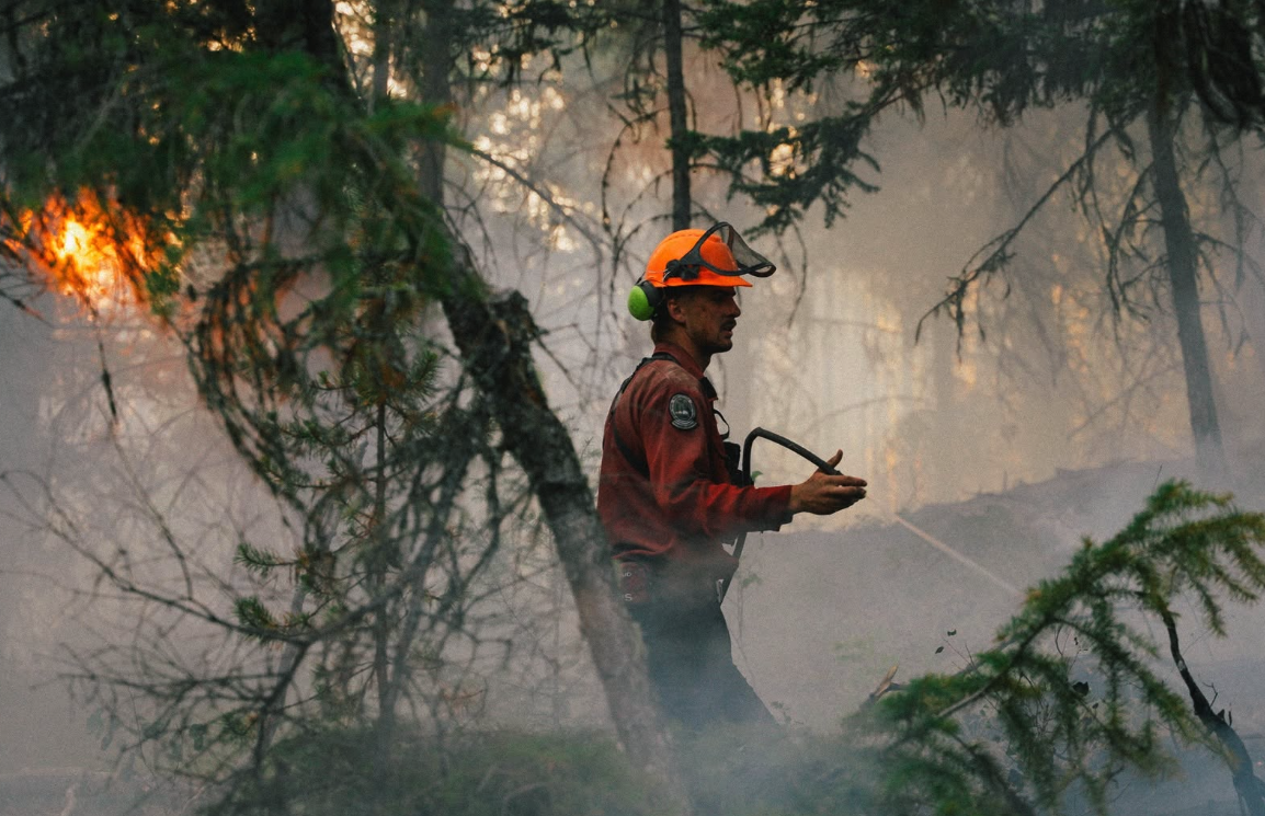 A BC Wildfire Service worker sprays a hose in a burning forest.