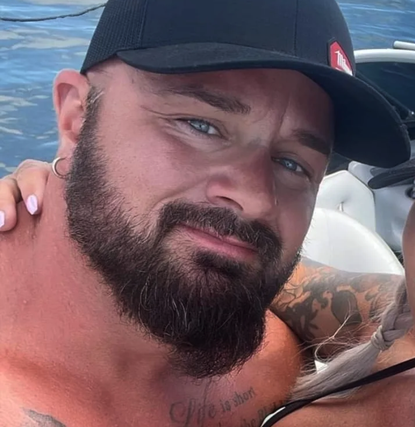A shirtless man with a beard wearing a baseball cap poses for a photo.