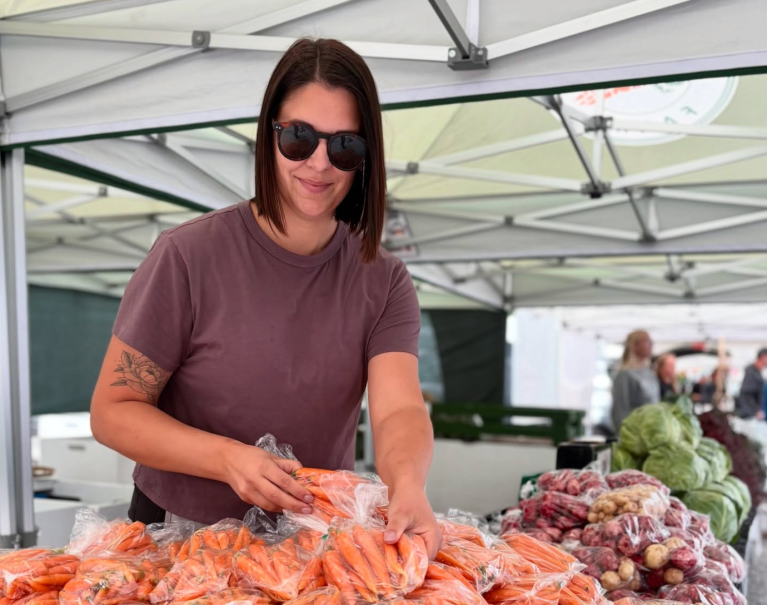 A woman wearing sunglasses stands behind a table with vegetables displayed.