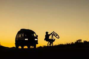 A van beside a person popping a wheelie on a bike are silhouetted against a soft, golden coloured sky.