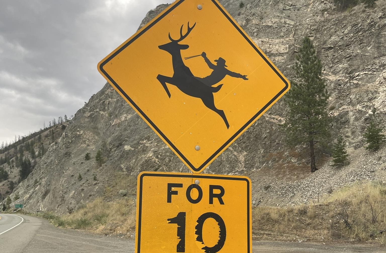 A yellow road sign on a highway shows the silhouette of a cowboy riding a deer.