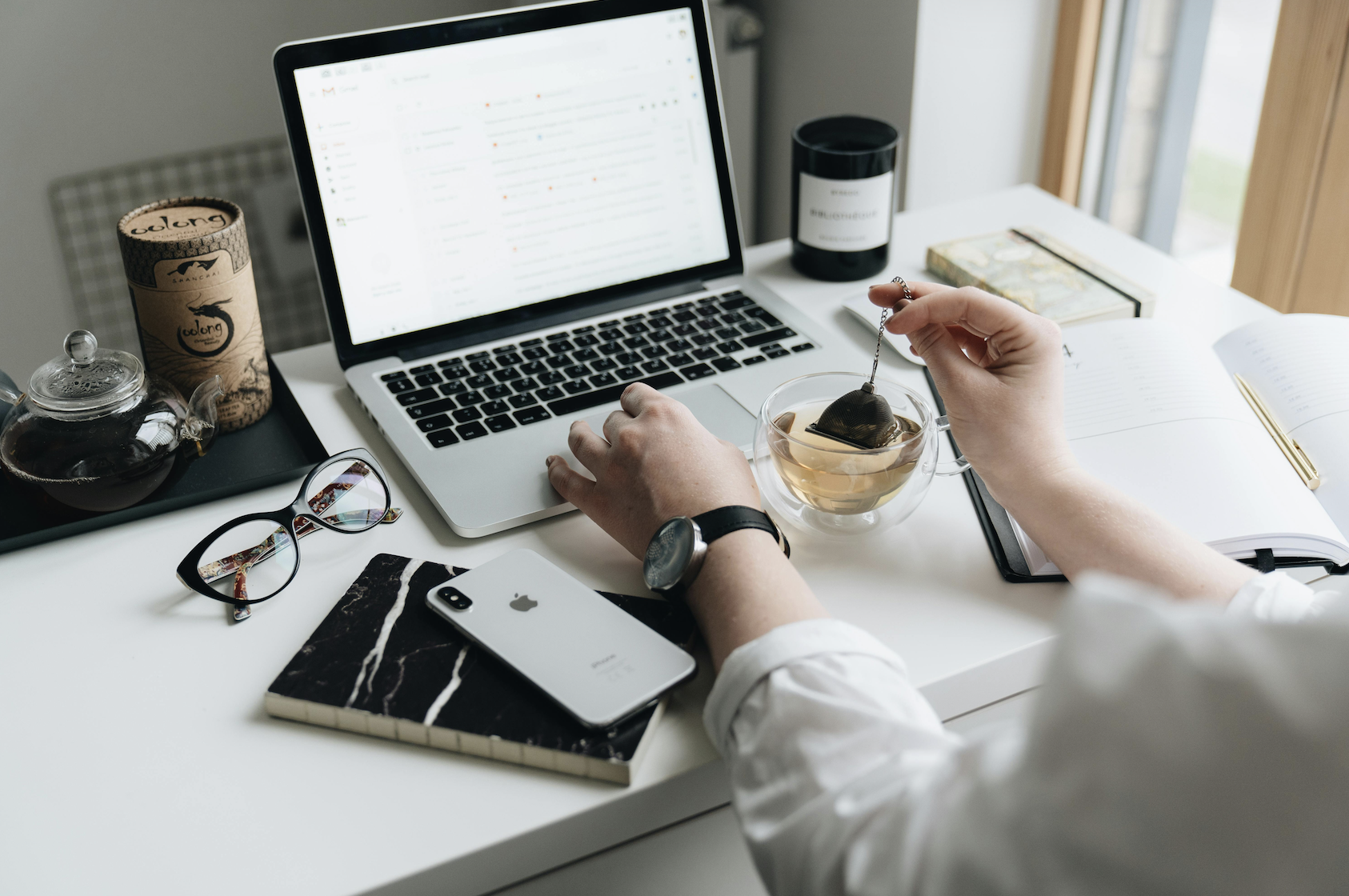 A woman at a desk looks at a laptop computer while making a cup of tea.