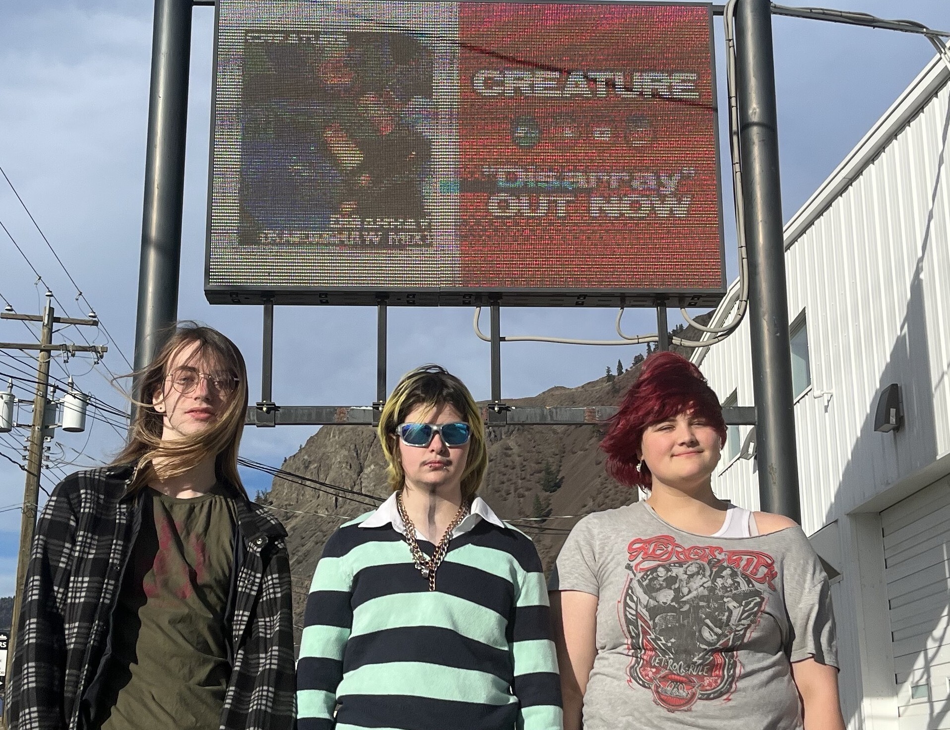 Three teenagers stand below a digital billboard.