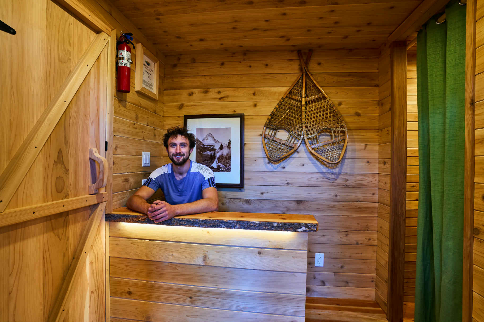 A man in a blue shirt smiles from behind the desk of a rustic cedar reception area.