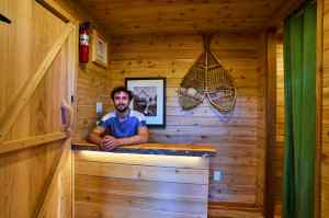 A man in a blue shirt smiles from behind the desk of a rustic cedar reception area.