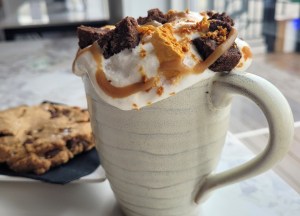 A mug of hot hot chocolate has candy, brownies and whipped cream on top of it.