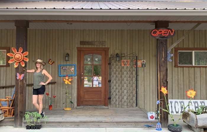 A woman wearing a cowboy hat, grey tank top and black shorts poses on the wooden step of a wooden store front that is decorated with metal art pieces.