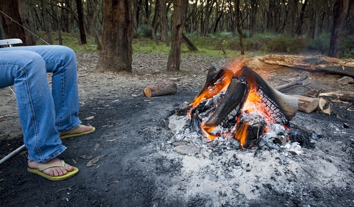 A mans sits on a lawn chair in front of a campfire.