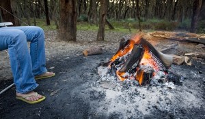 A mans sits on a lawn chair in front of a campfire.