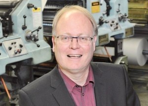 A man poses for a photo in front of a newspaper printing press.