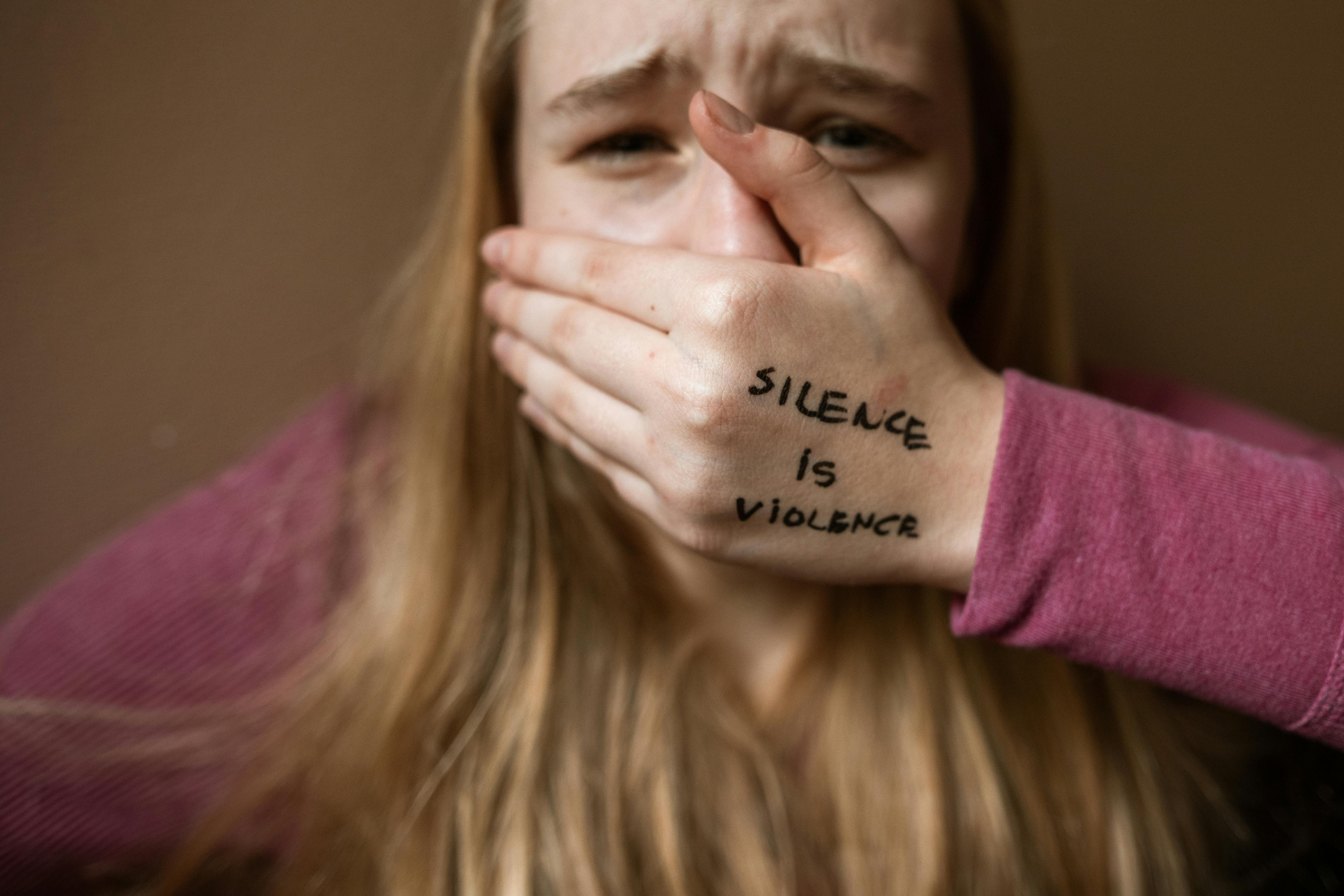 An upset woman with the words Silence is Violence written on the back of her hand which is covering her mouth.