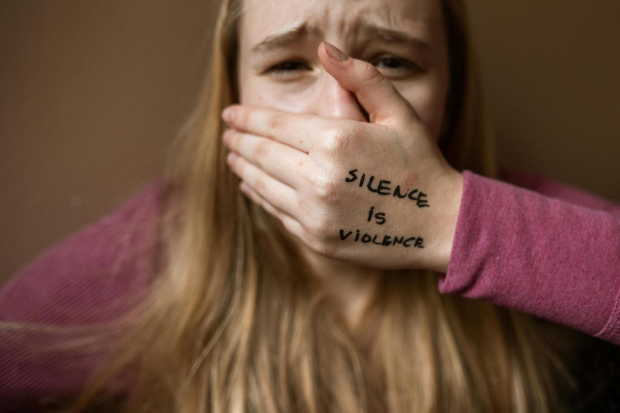 An upset woman with the words Silence is Violence written on the back of her hand which is covering her mouth.