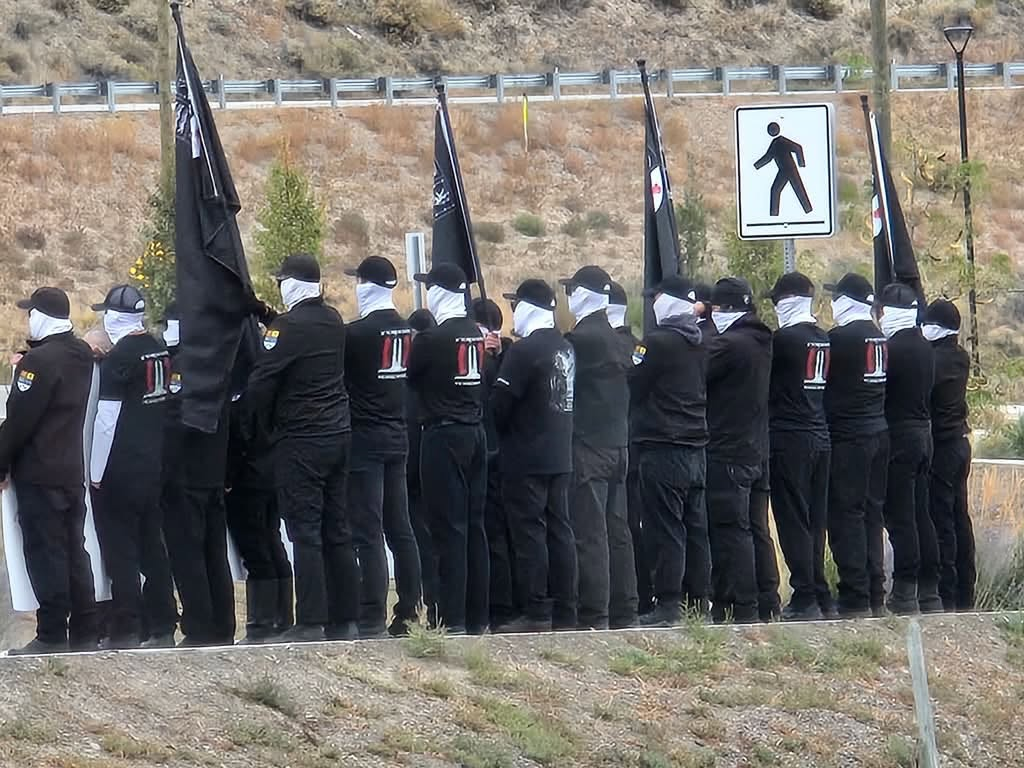 People in a line with their backs turned wearing black clothes and white face coverings.