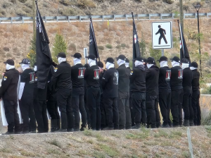 People in a line with their backs turned wearing black clothes and white face coverings.