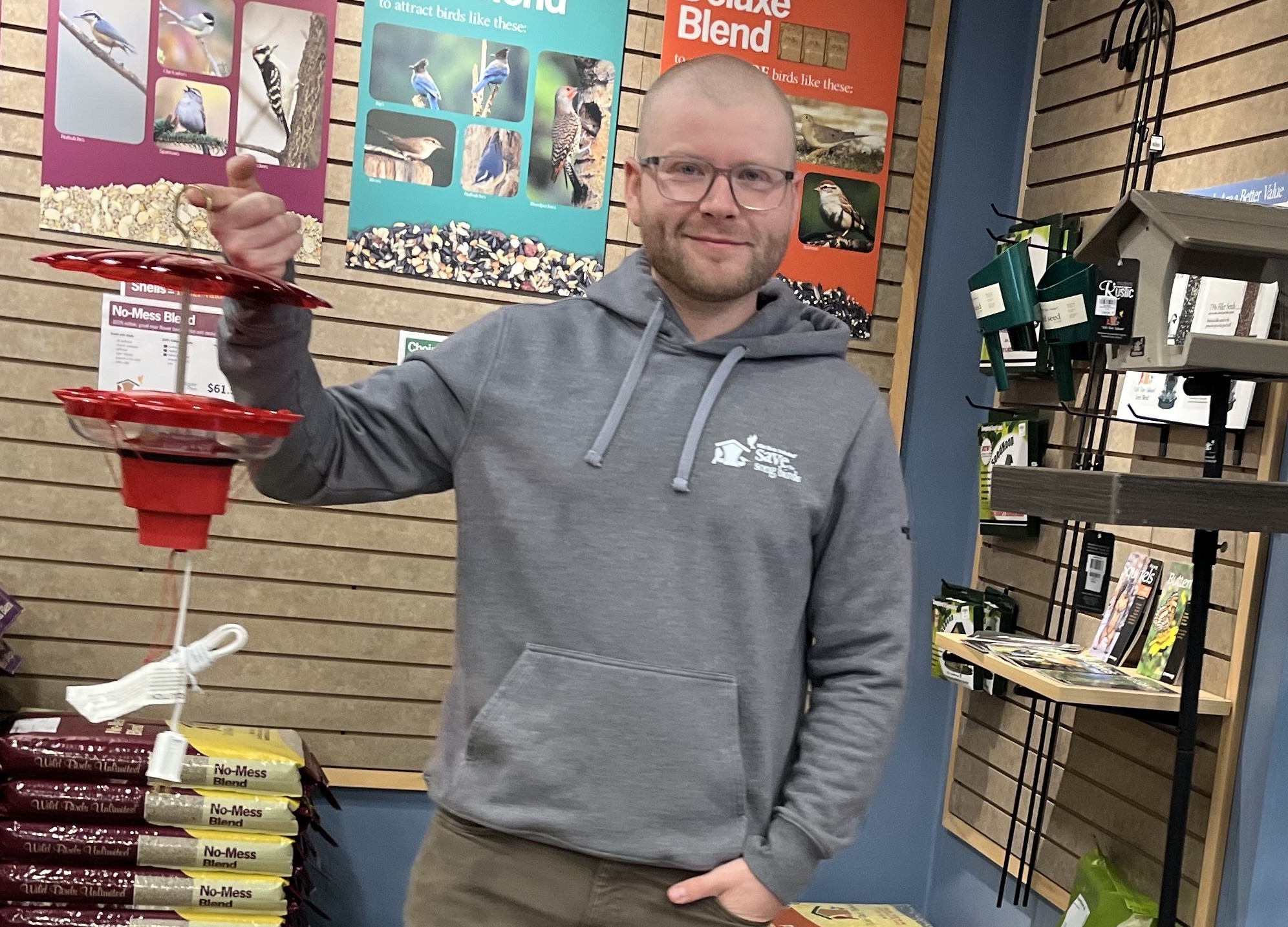 A man holds a hummingbird feeder up with his left hand while his right hand is in his jeans pocket.