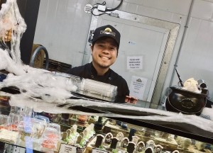 A smiling man in a black hat stands behind a bakery display.