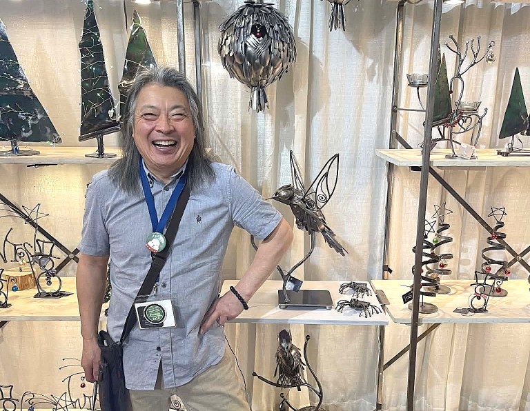A man stands with his hand on his hip wearing a smile in front of metal sculptures.