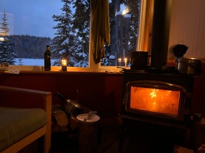 The inside of a cabin is dimly lit with a burning stove in the corner and window.