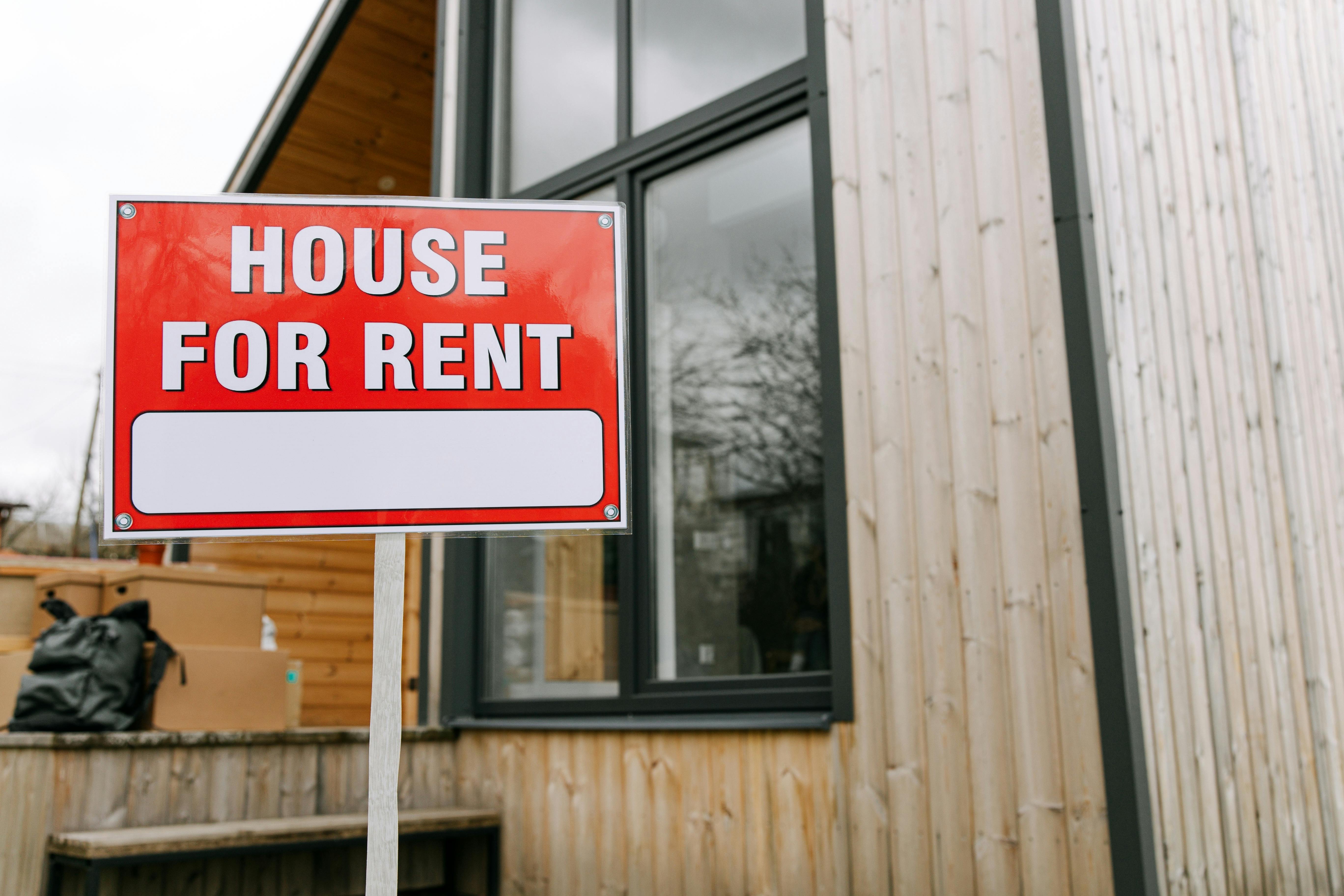 A red 'house for rent' sign in front of a woodsided property,