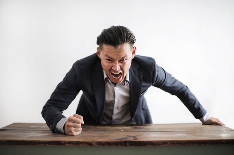 A man in a suit pounds a table with his fist in a fit of rage.