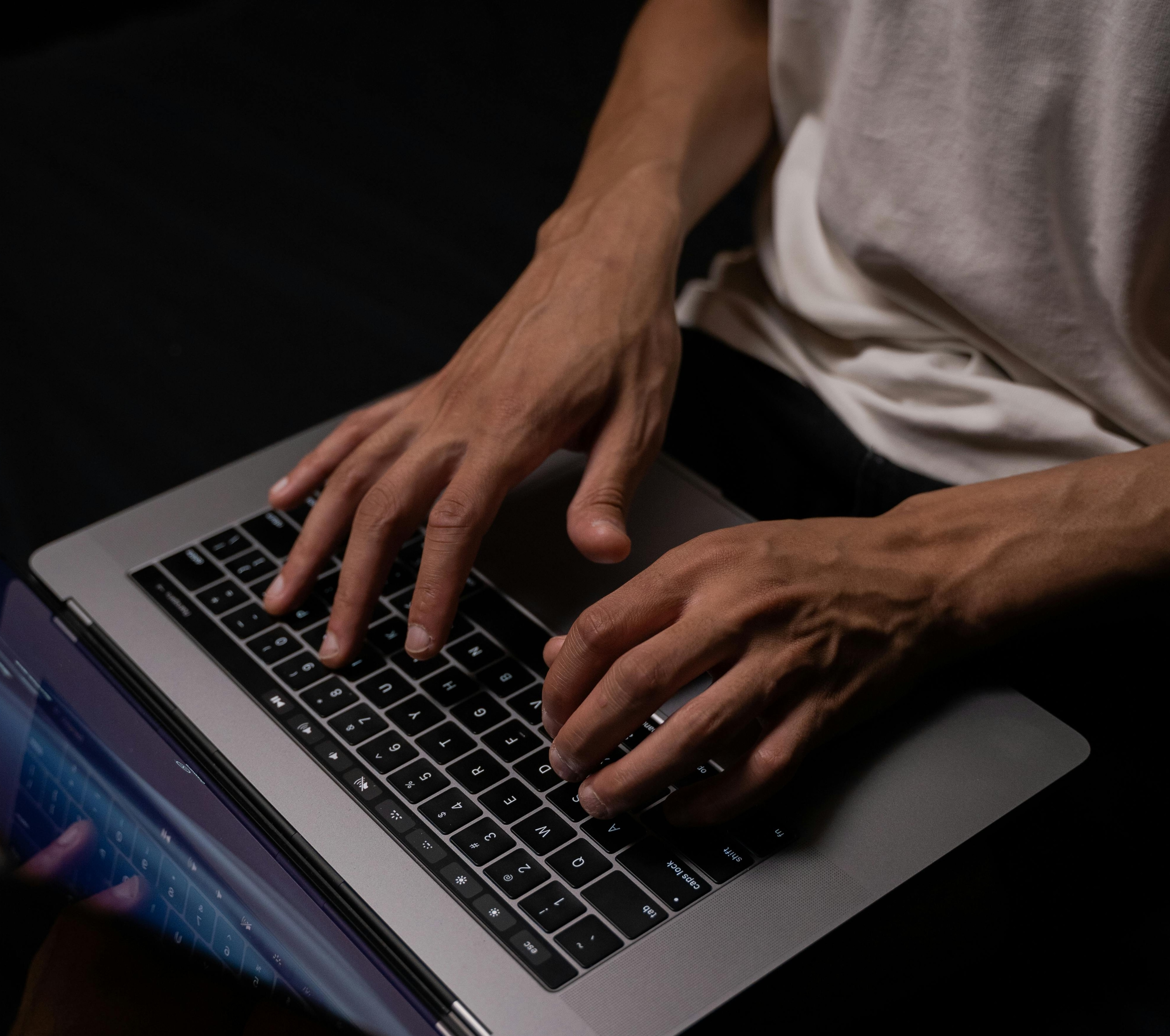 A man types on a keyboard of a laptop computer in the dark.