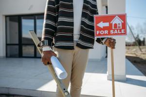 A man stands with a roll of paper and a for sale sign outside a house.
