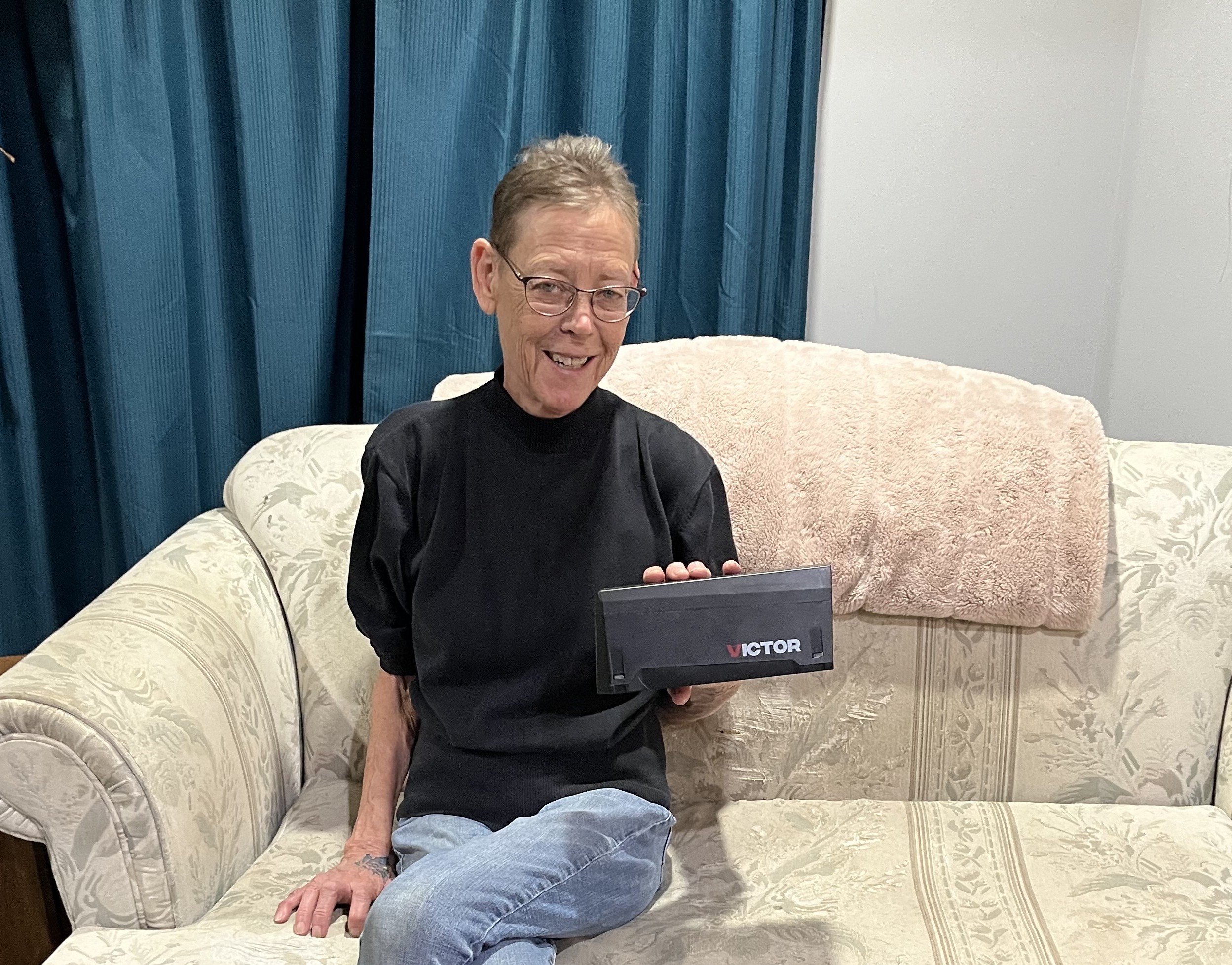 A woman sitting on a sofa holds up a rectangular black rat trap.