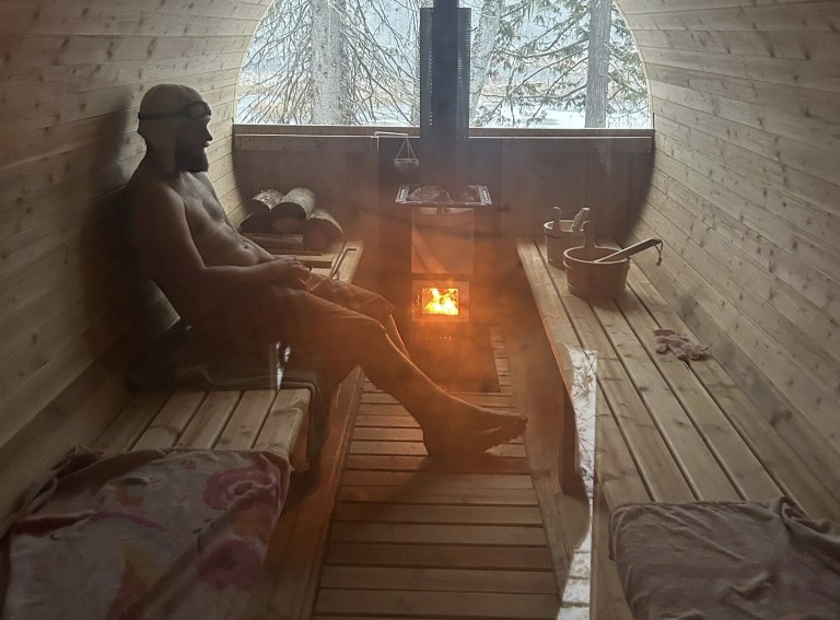 A man sits by a stove in an outdoor sauna.