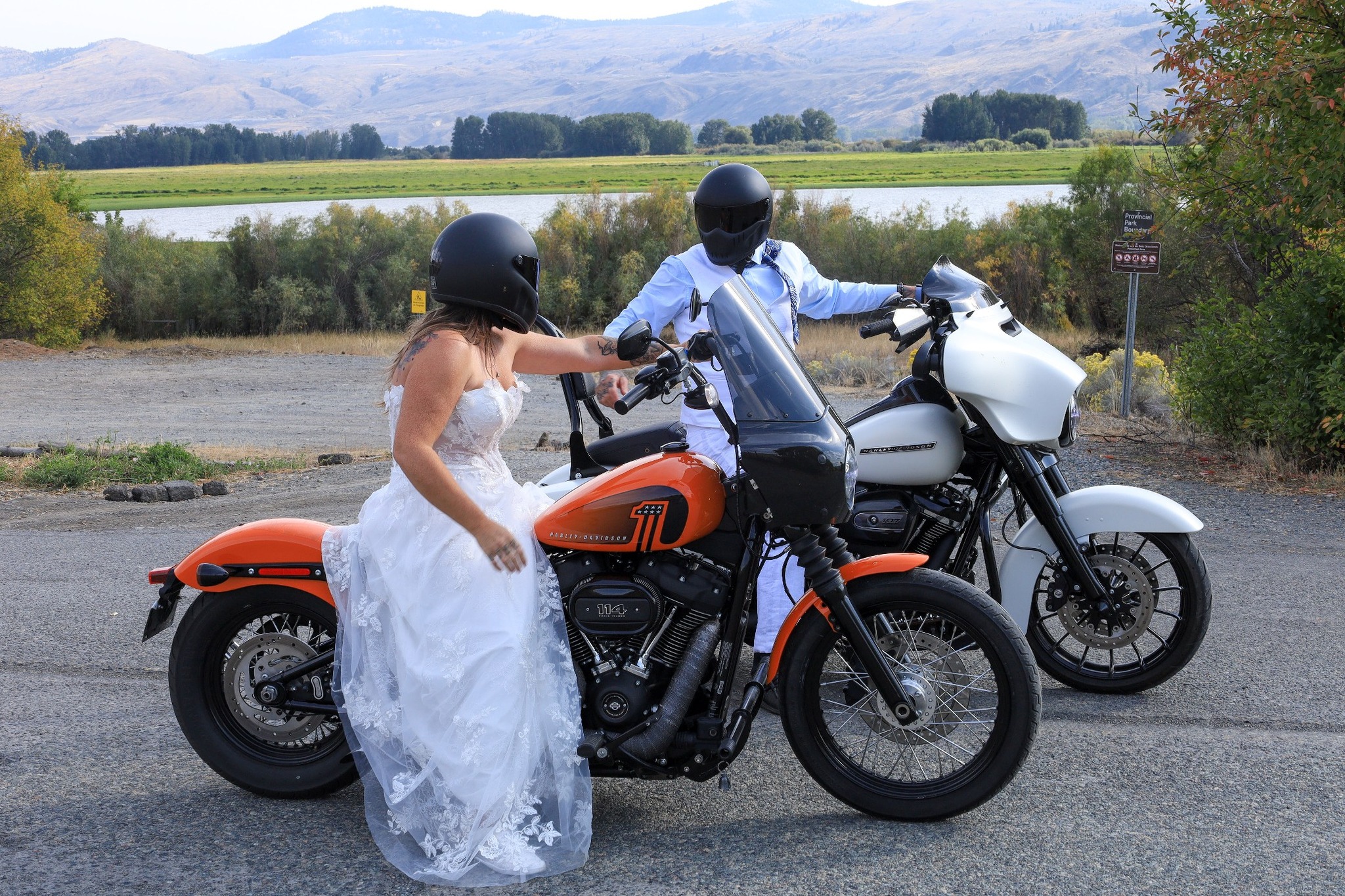 I woman in a white wedding dress sits on an orange motorcycle in the foreground with a man in a blue shirt sits in a white motorcycle behind her.